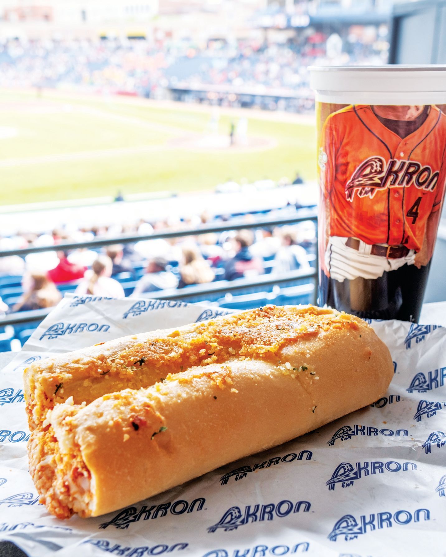A stadium snack of a long sub sandwich and a branded cup, overlooking a baseball game from the stands.