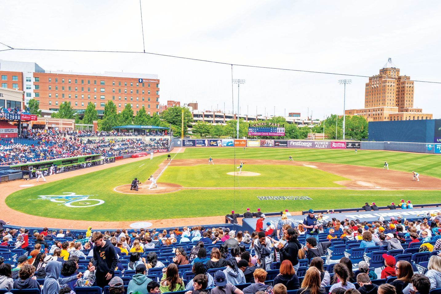 A sunny day at a minor league baseball stadium with spectators in the foreground and a tan building in the background.