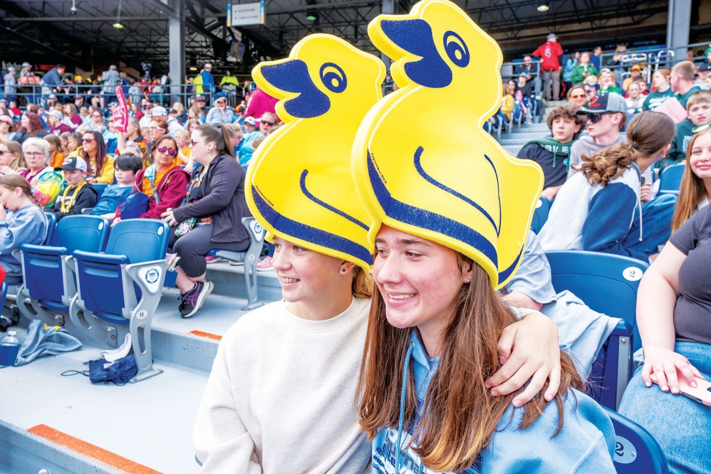 Two people in stadium seats wearing large, yellow duck-shaped hats and smiling at a sports event.