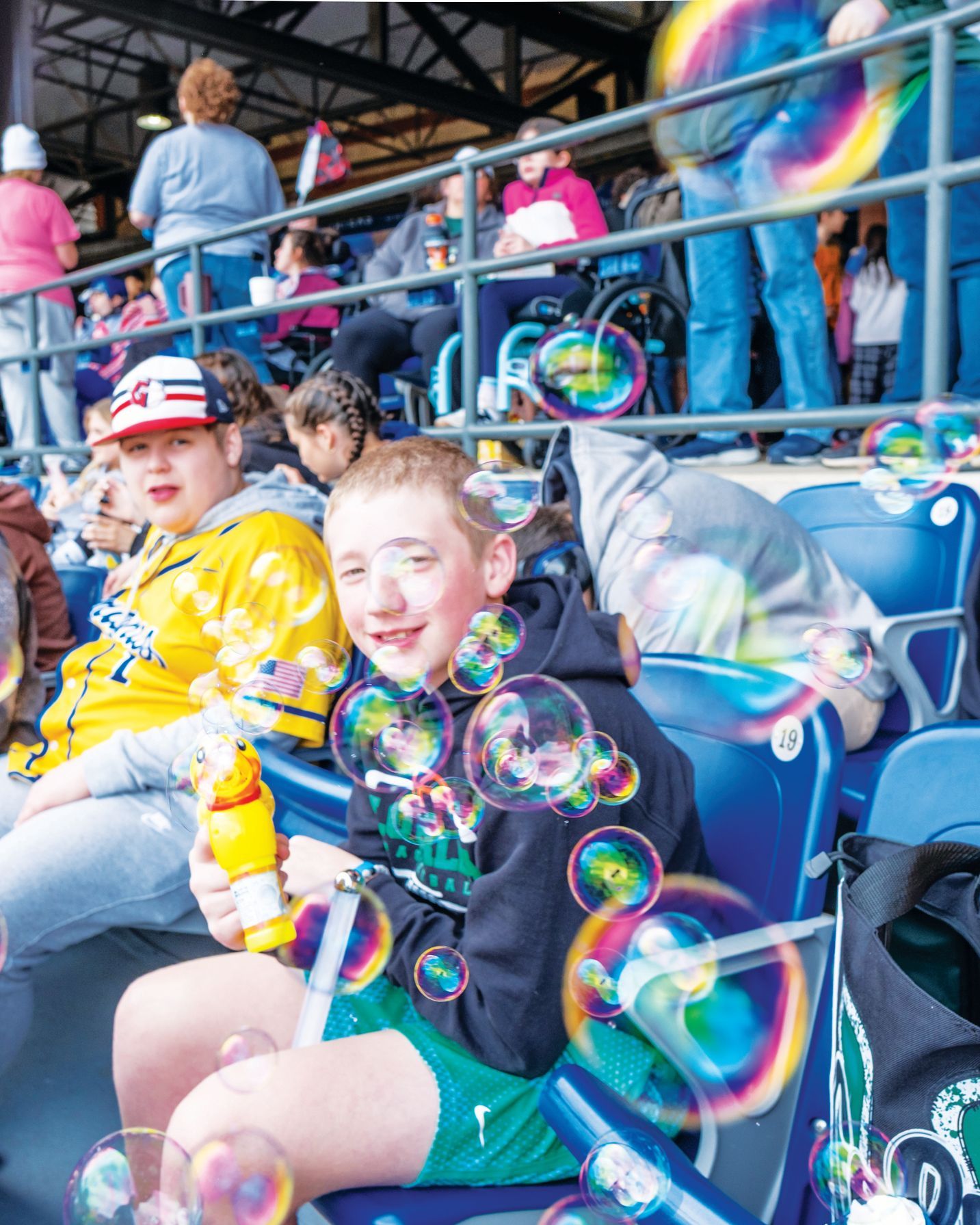 People in stadium seating watch as bubbles float in the air in front of a smiling person holding a yellow bubble wand.