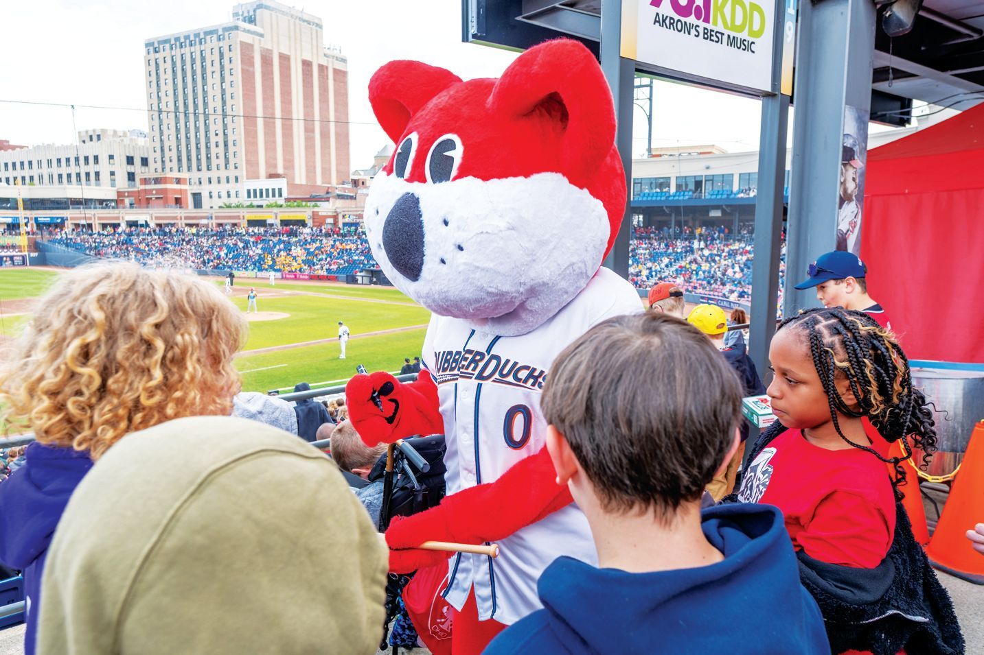 A mascot with red and white fur interacts with a group of people at a baseball stadium.