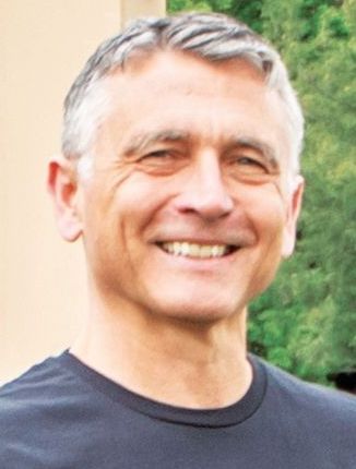 A smiling person with short grey hair, wearing a dark blue t-shirt against a blurred outdoor background.