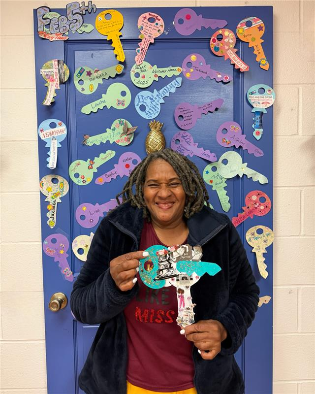 A smiling individual holds a decorated key cutout in front of a blue door covered in numerous handmade paper keys.