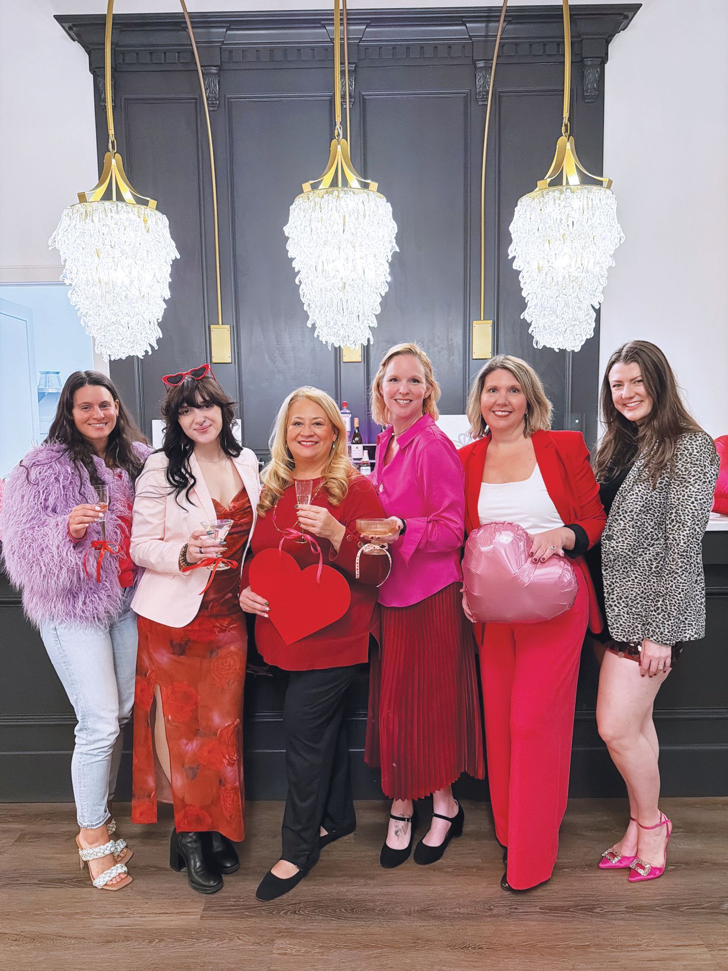 Six people pose together at a celebration, wearing red, pink, and festive clothing, holding hearts in an indoor setting.