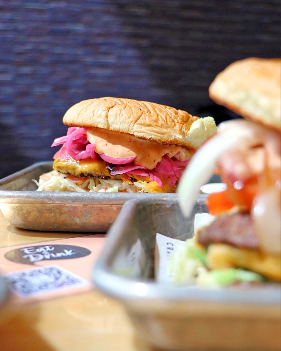 Two burgers with pickled onions and sauce on metal trays, with a blurred foreground and an
