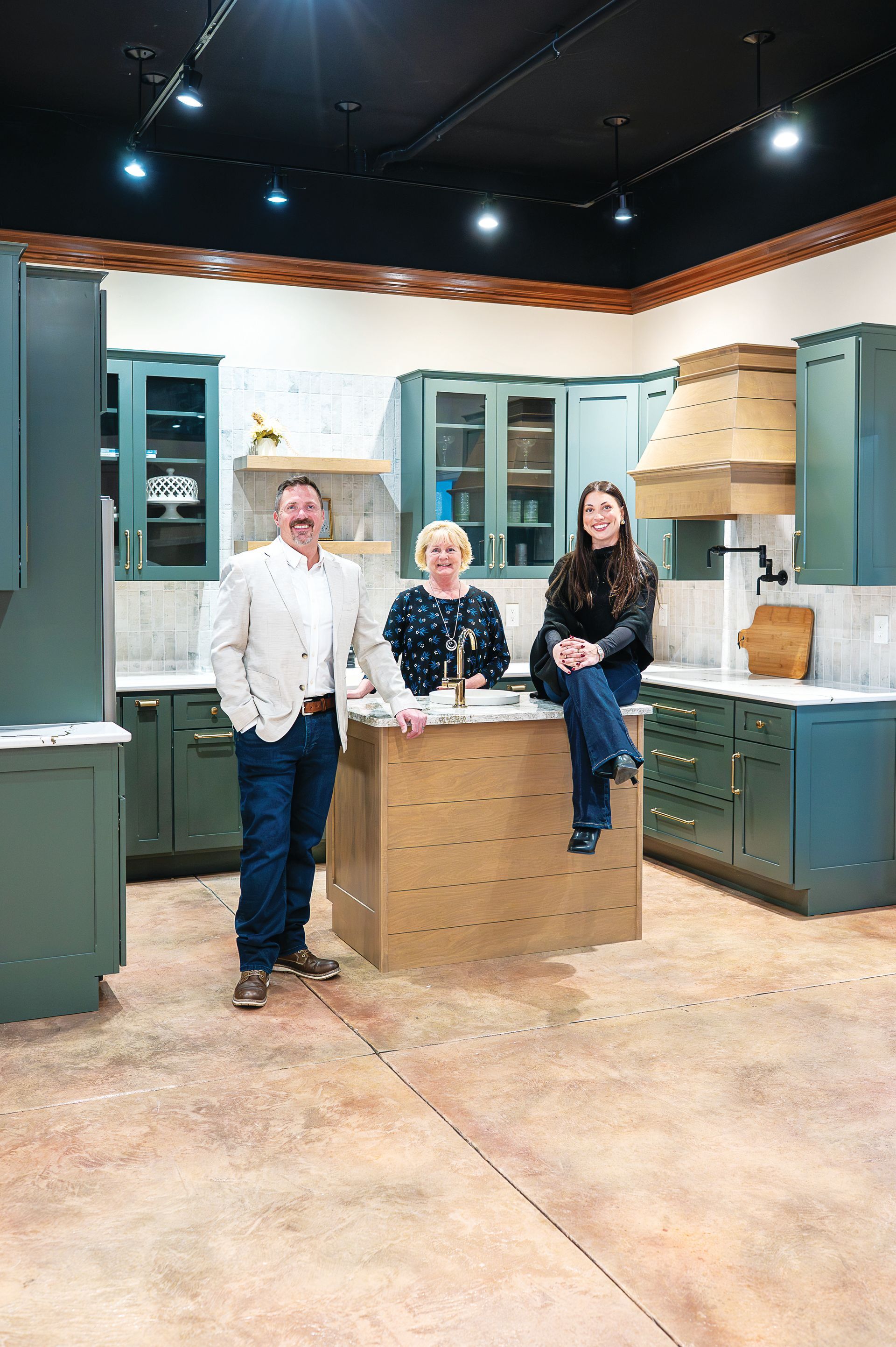 Three people stand and sit in a kitchen showroom with green cabinets, a light wood island, and a textured concrete floor.