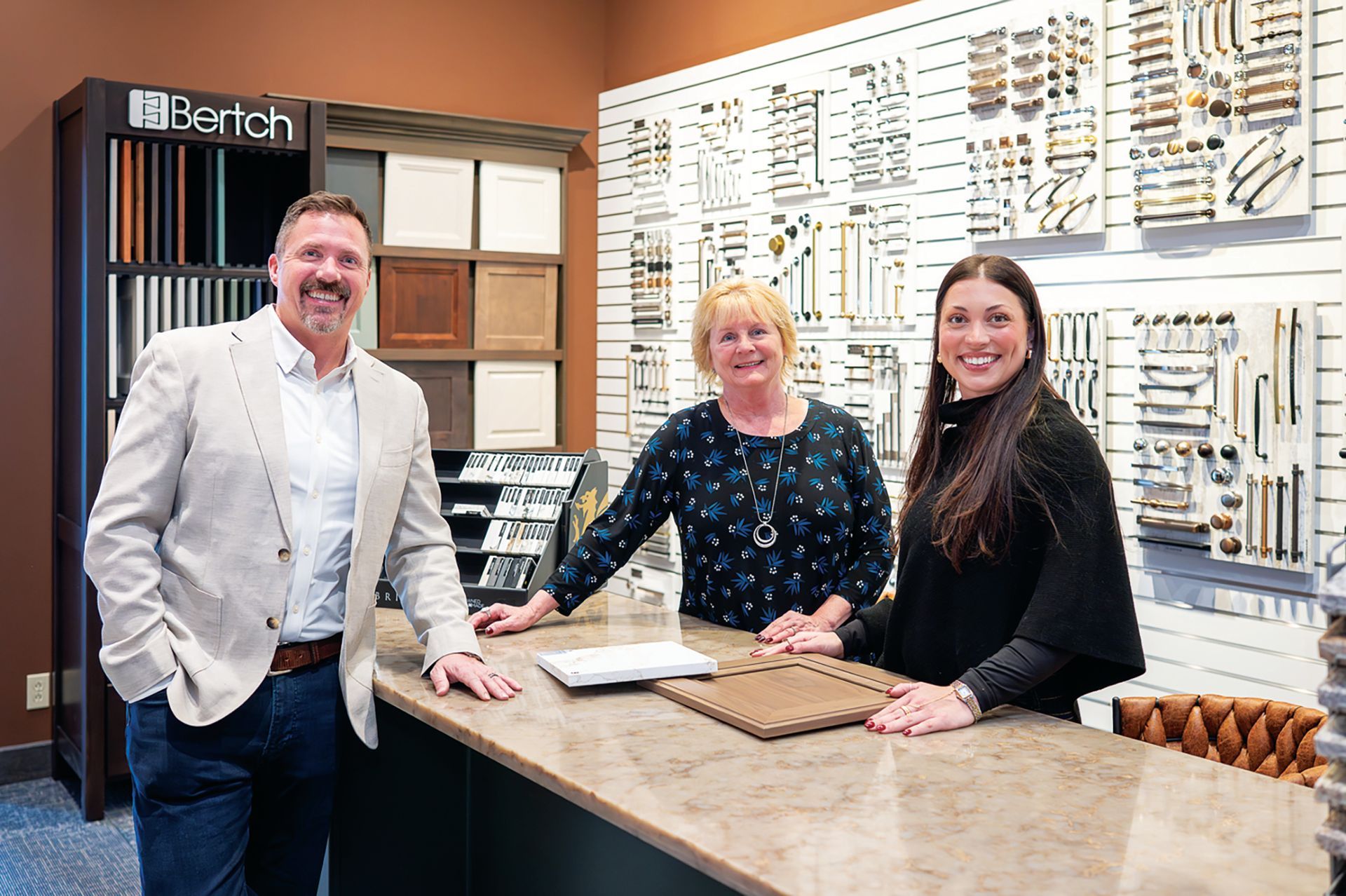 Three professionals smile behind a countertop in a showroom filled with cabinet and hardware samples.
