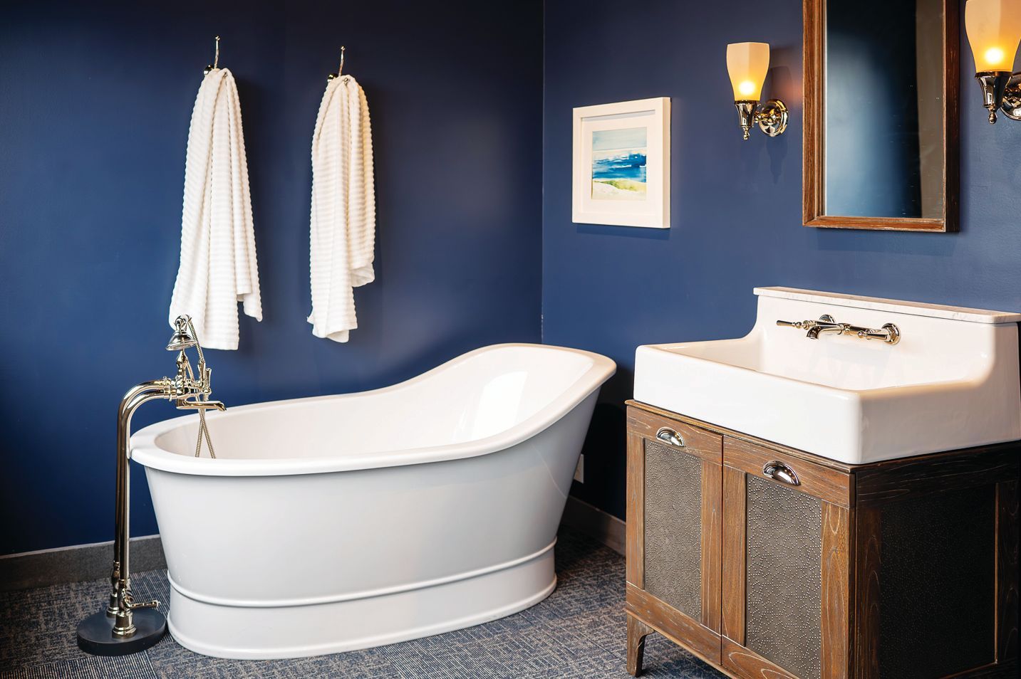 A bathroom with navy walls, a white freestanding soaking tub, a farmhouse-style sink on a wooden vanity, and wall art.