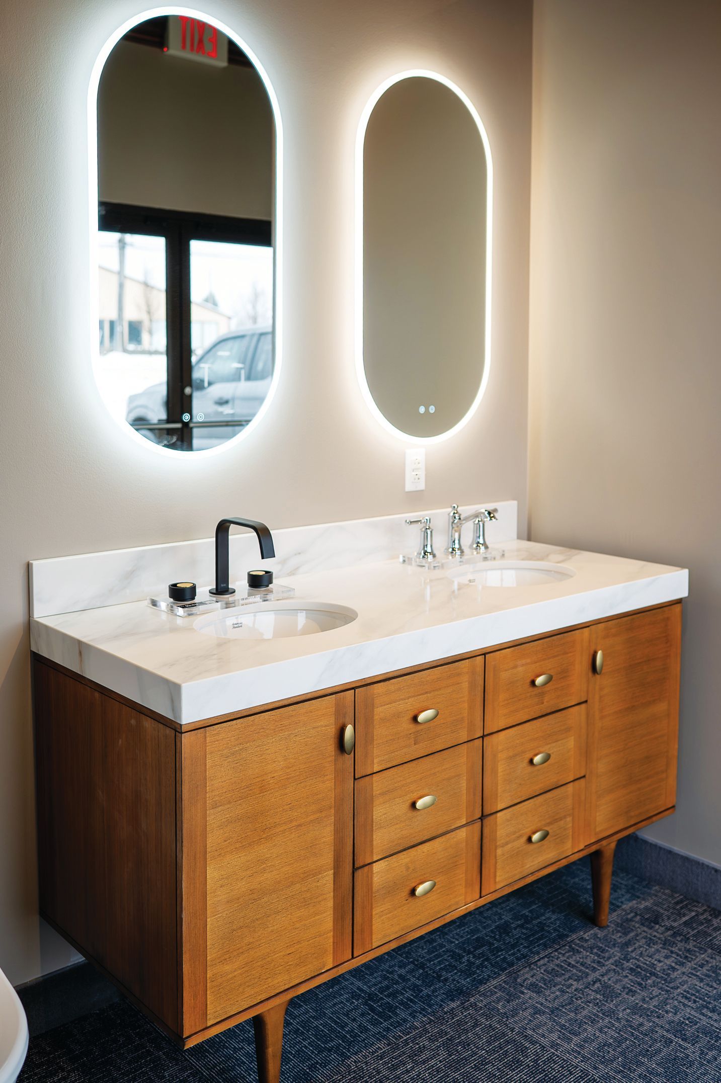 A double-sink bathroom vanity with wood cabinetry, marble countertop, and two oval backlit mirrors on a beige wall.