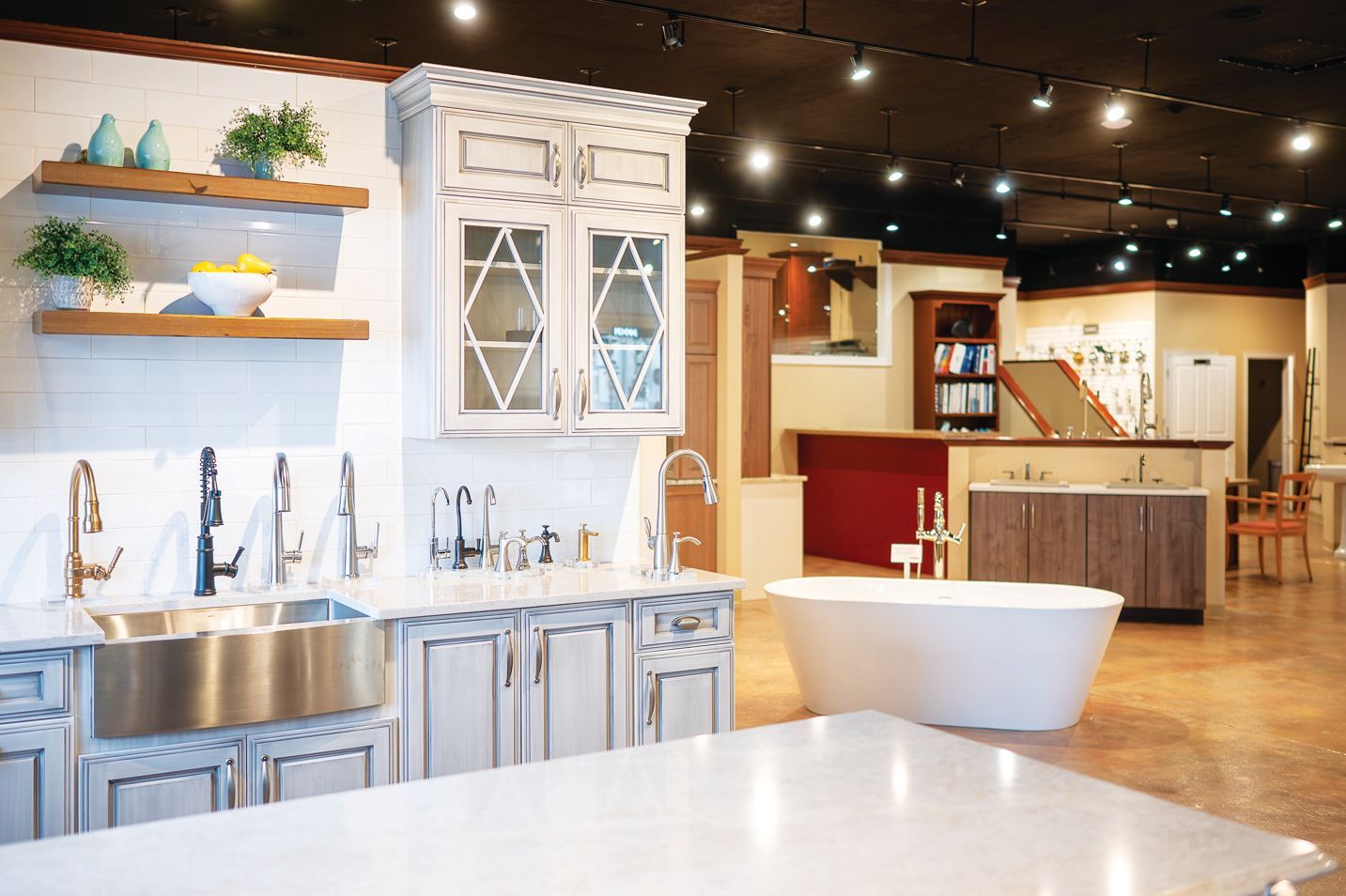 A showroom display featuring white kitchen cabinets, a farmhouse sink, shelving, and a freestanding soaking bathtub.