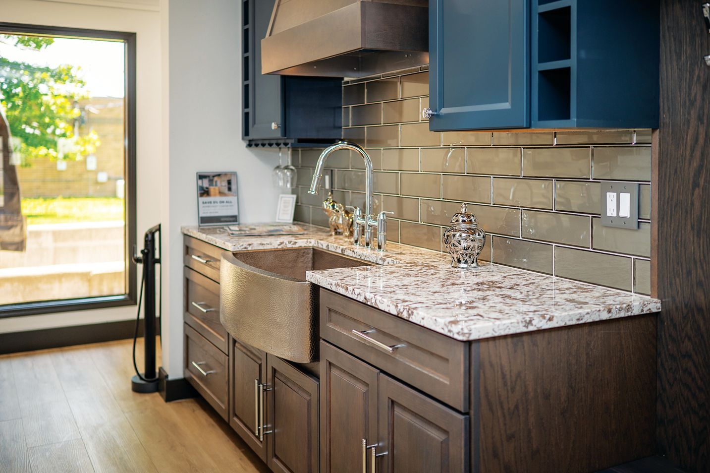 A kitchen corner with brown wood cabinets, a hammered copper farmhouse sink, white speckled counters, and blue cupboards.