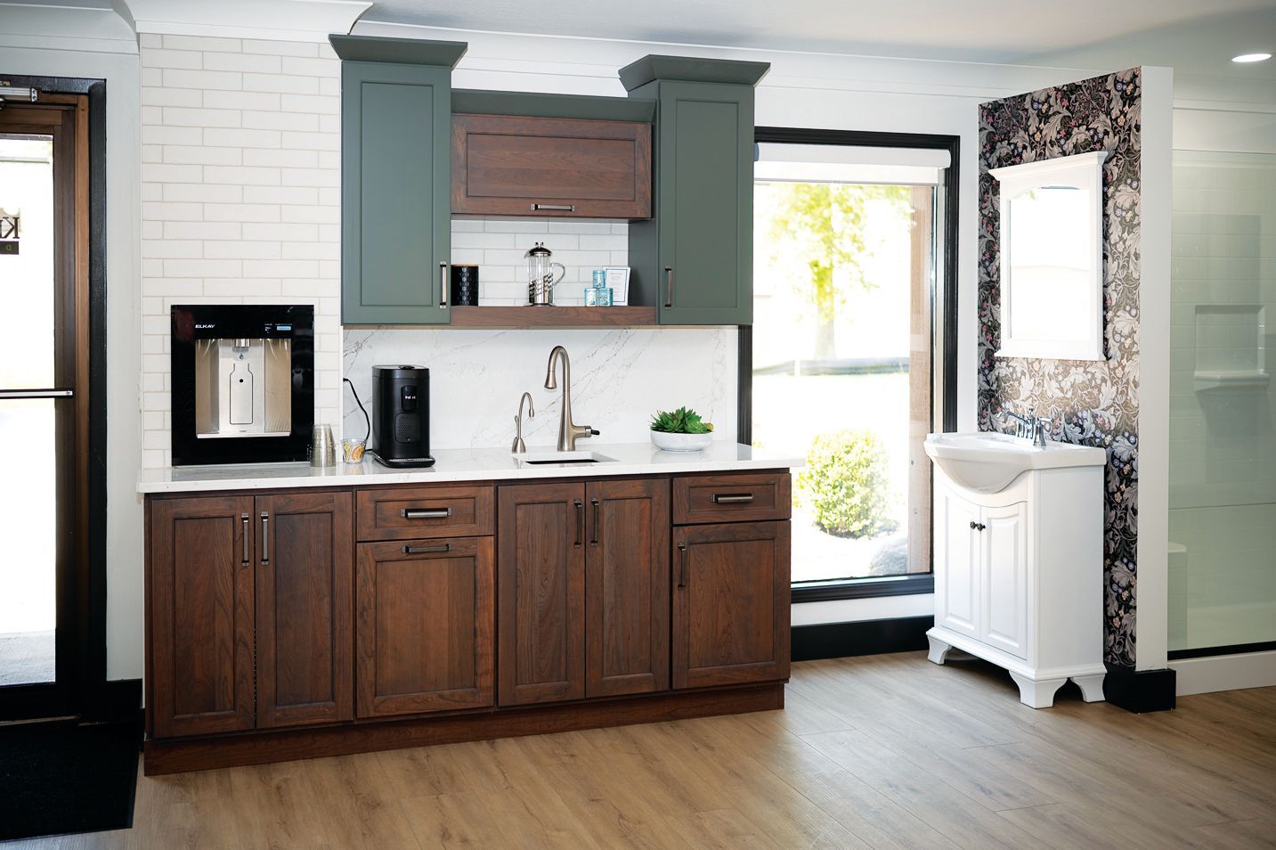 A modern kitchenette with dark wood cabinets, green upper cupboards, white backsplash, and a separate white vanity.