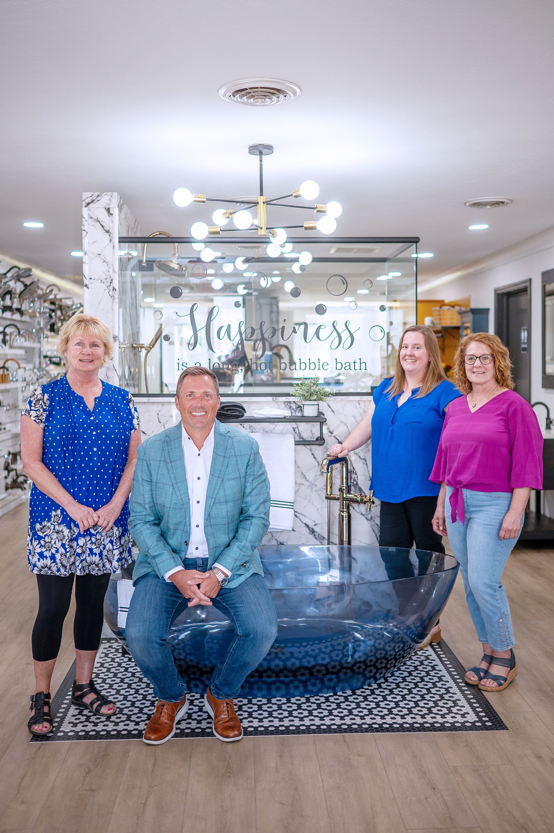 Four people stand in a showroom, one sitting on a blue bathtub, posing in front of a mirror with a decorative chandelier.