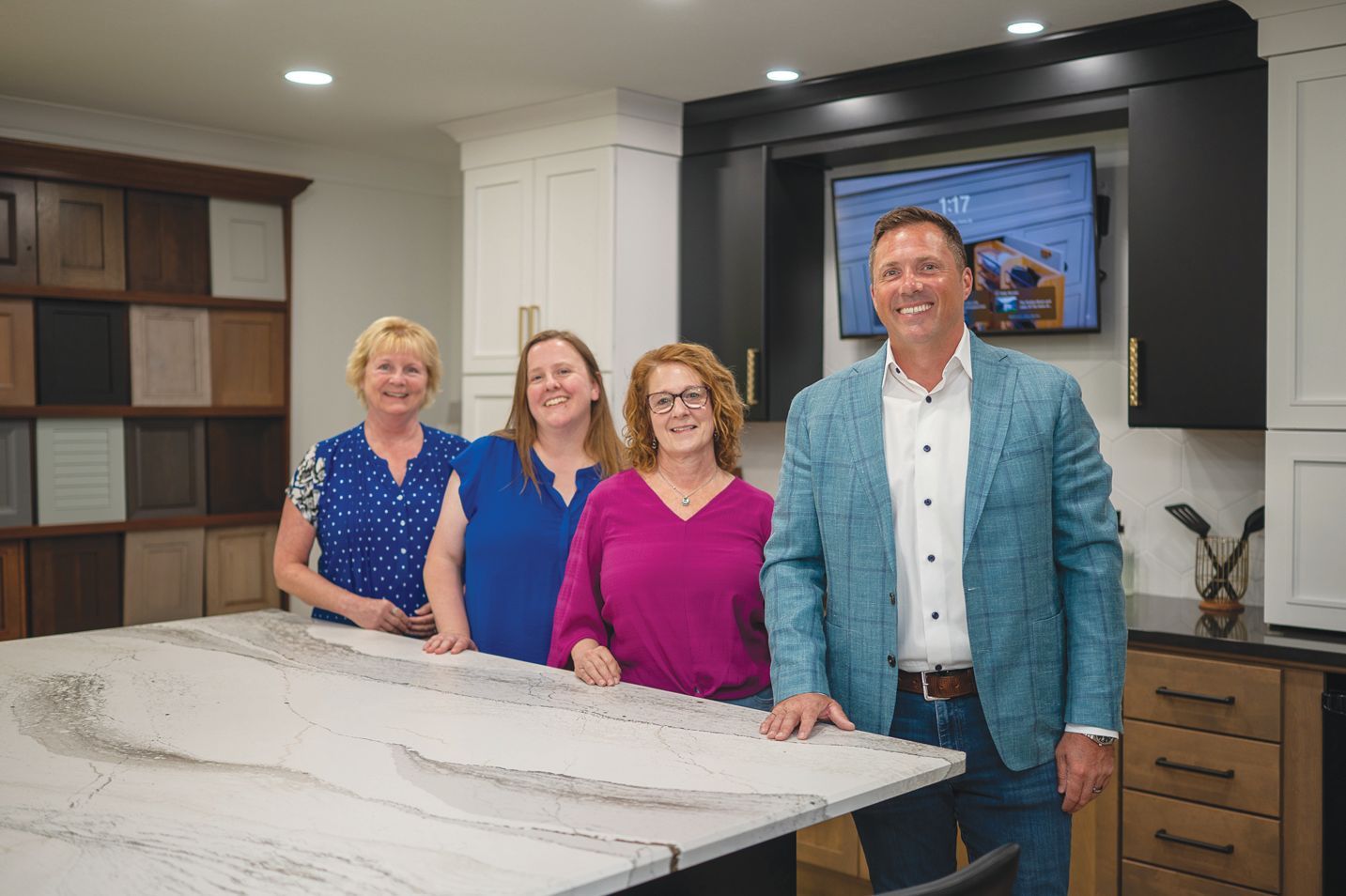 Four smiling professionals stand behind a large stone kitchen island in a showroom featuring various cabinet samples.