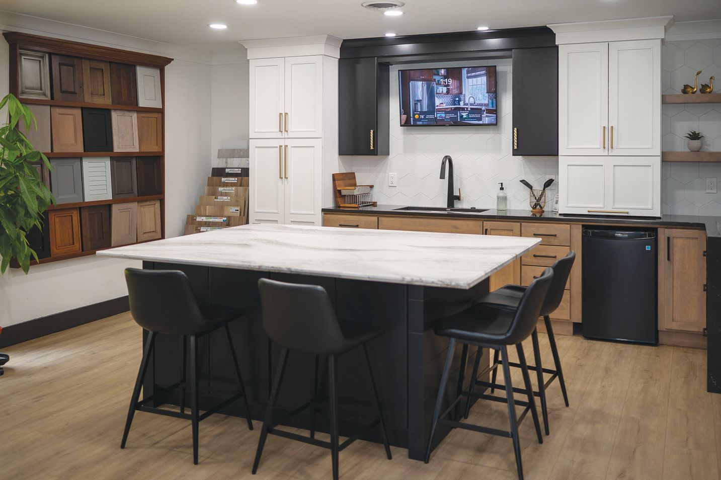 A showroom featuring a kitchen island with black bar stools, a marble countertop, white and black cabinets, and a sink.