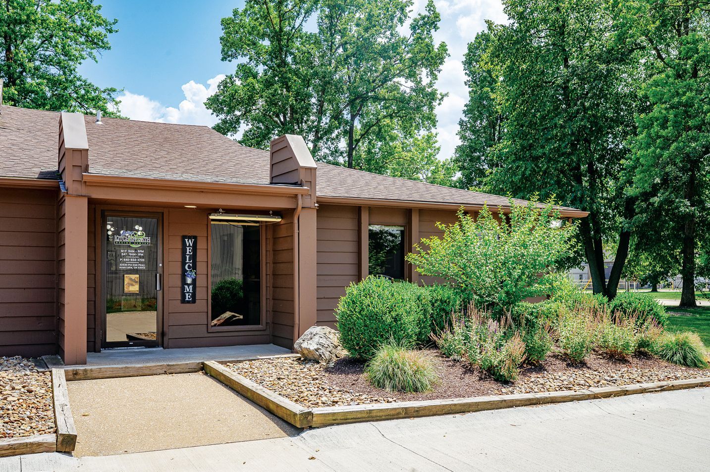 A wood-paneled building with a glass entrance, gravel path, and landscaping under a blue sky.