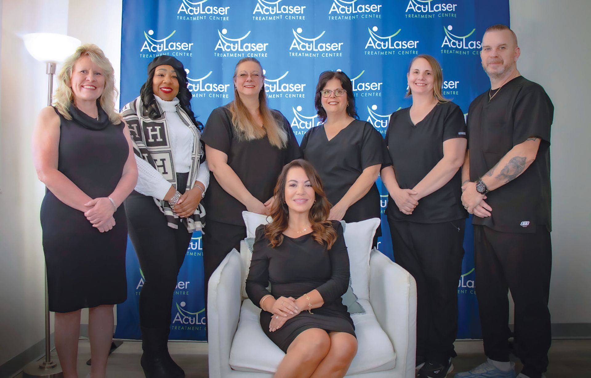 A group of seven people posed in front of a blue brand backdrop, with one person seated in a white chair.