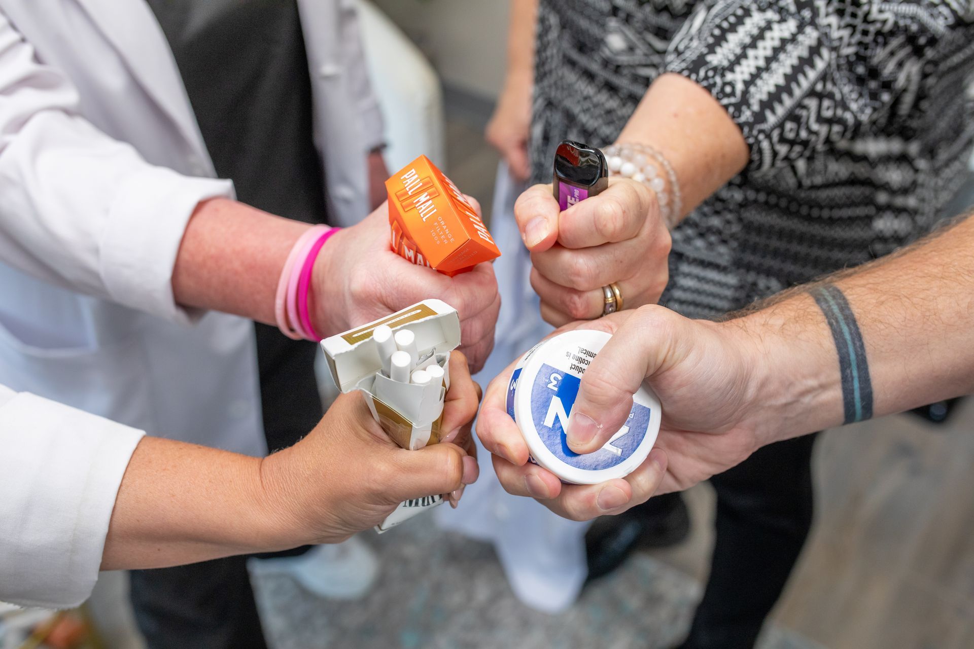 Multiple hands holding a pack of cigarettes, a lighter, a small container, and a package of nicotine replacement gum.