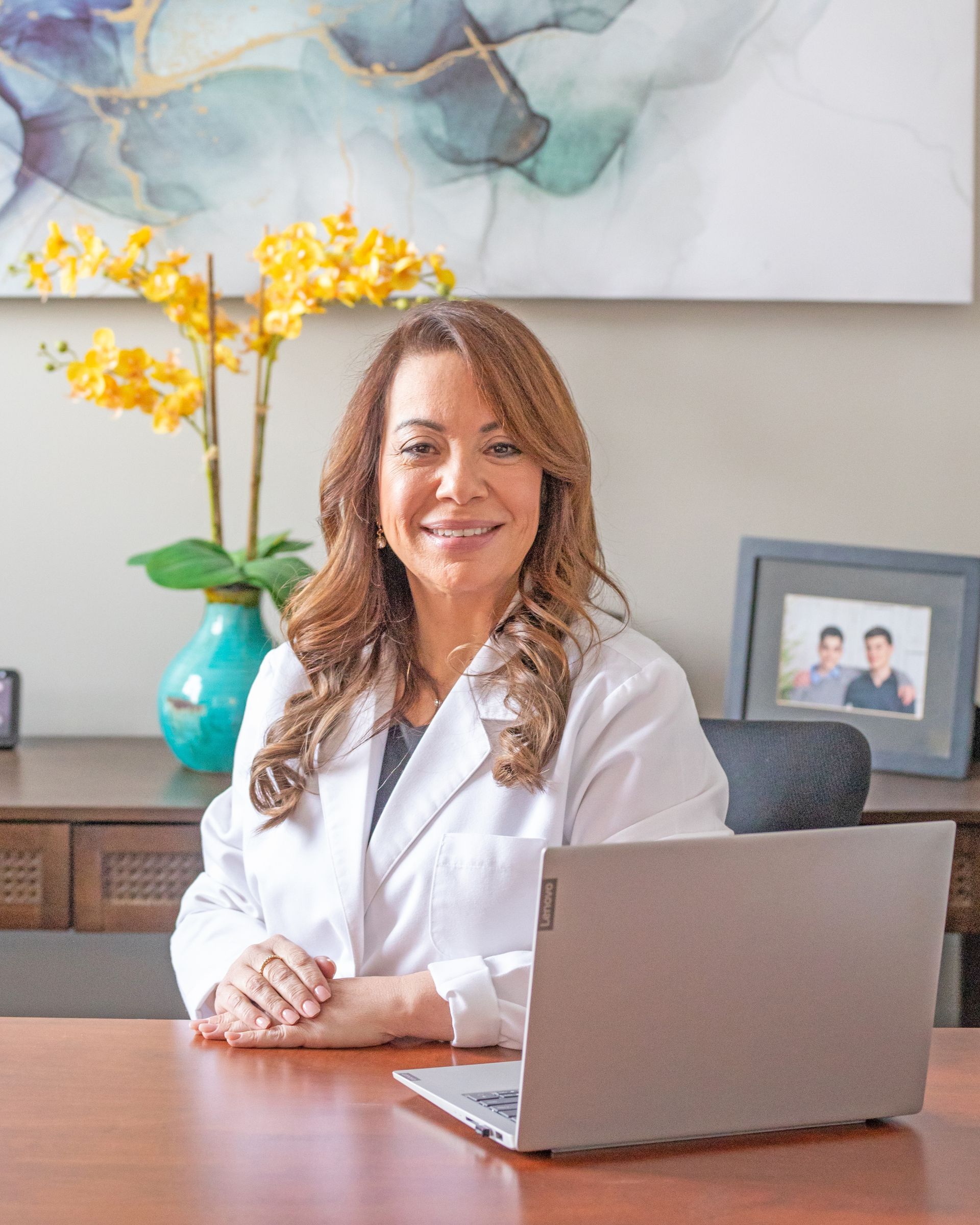 A professional sitting at a desk with a laptop, in an office with yellow flowers and framed photos in the background.
