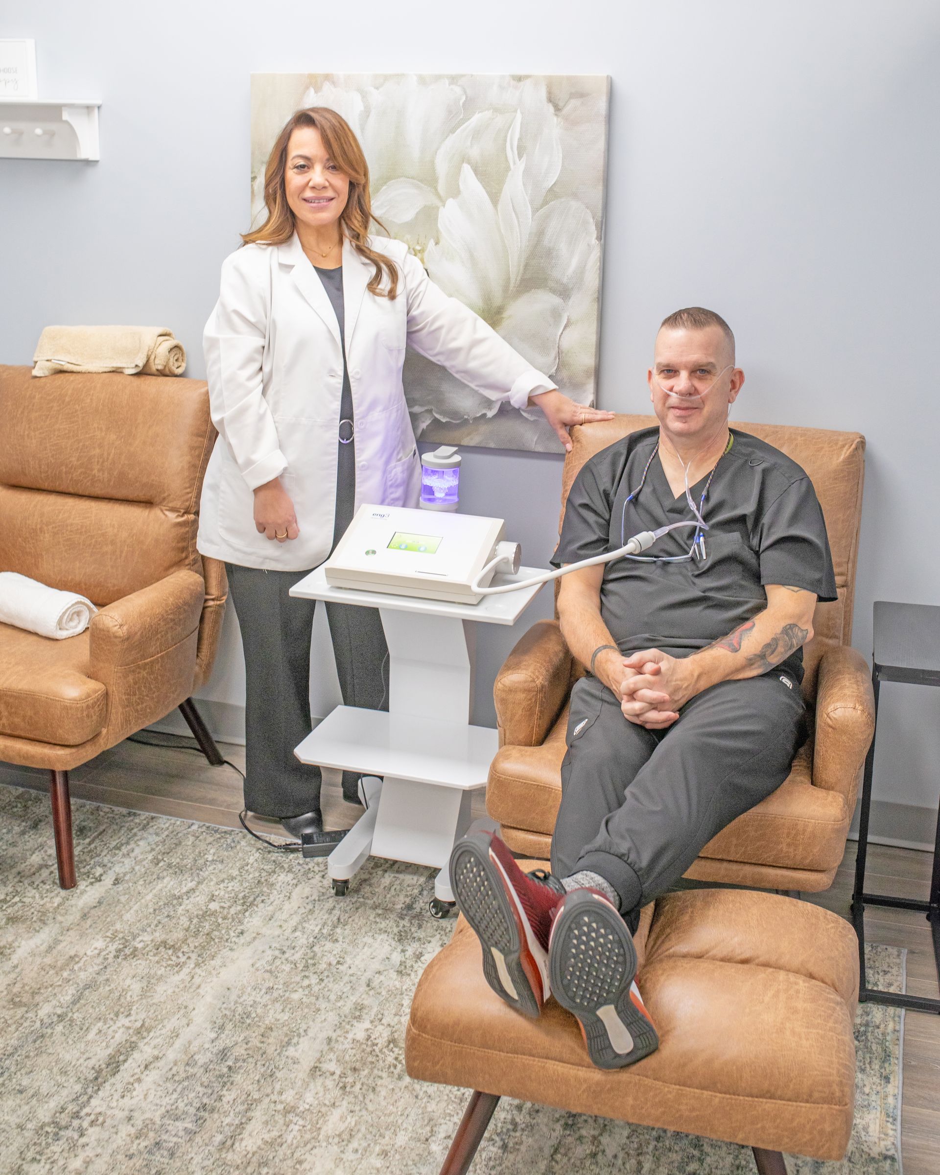 A medical professional stands next to a seated person connected to medical equipment in a neutral, modern office setting.