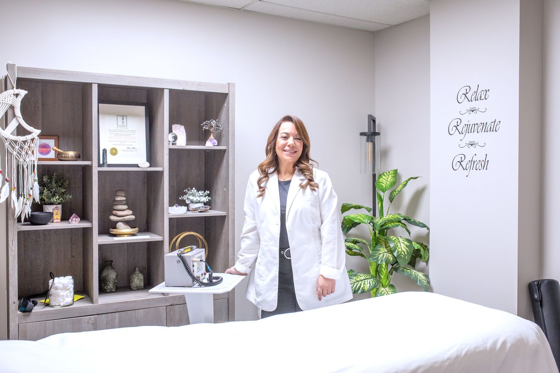 A professional in a white coat stands next to a treatment table in a wellness room with a shelving unit and a plant.