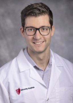 A professional headshot of a person in a white lab coat with a University Hospitals logo against a grey background.