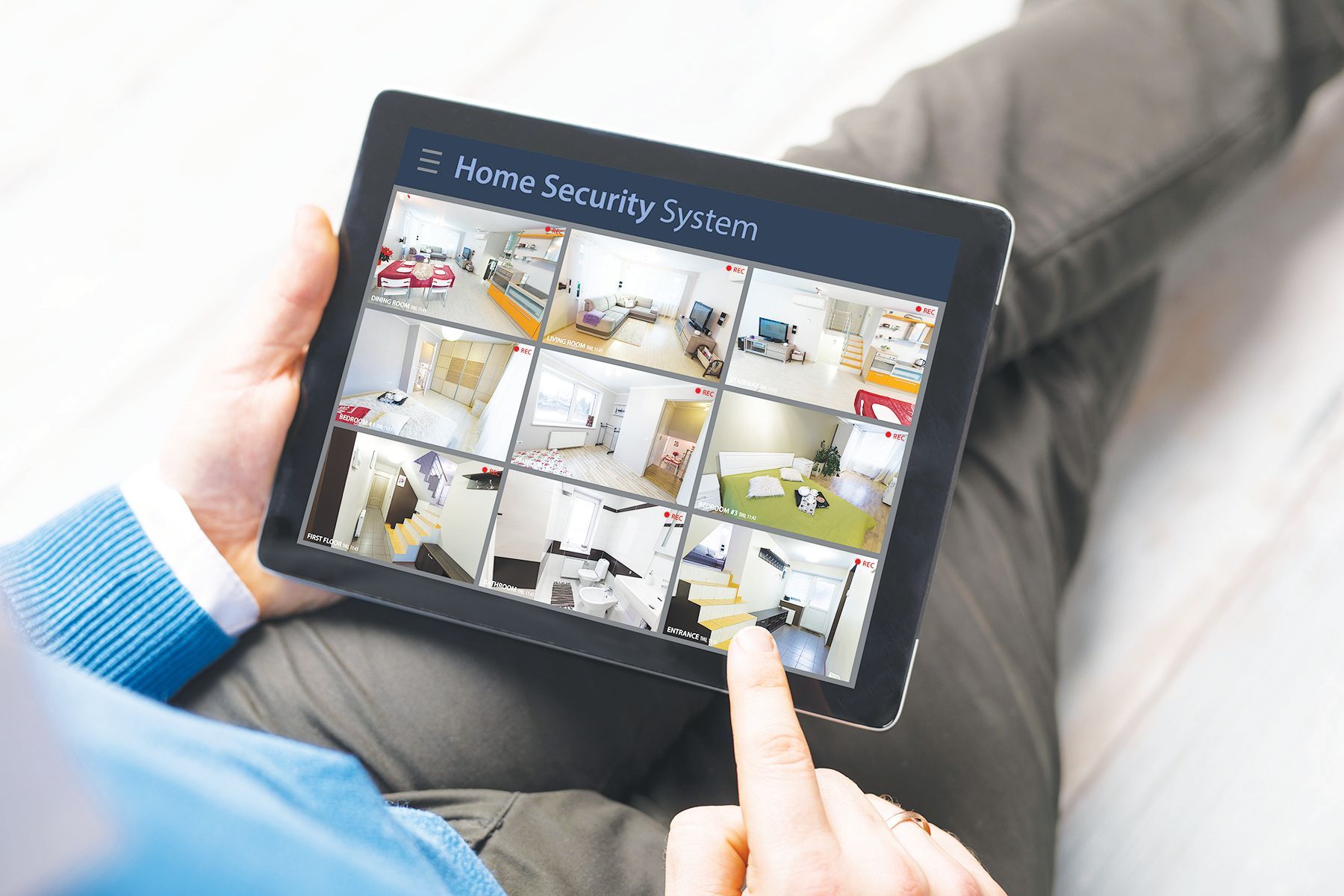A person in a blue sweater uses a finger to touch a tablet displaying a nine-camera home security system grid.