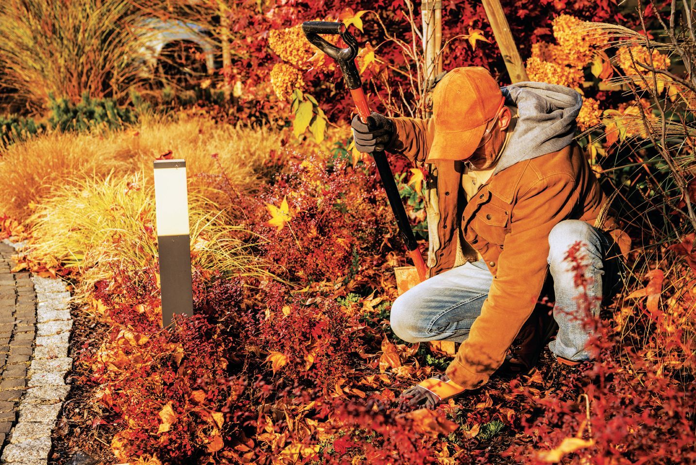 A person in a tan jacket and orange cap kneels in a garden among autumn leaves, holding a shovel.