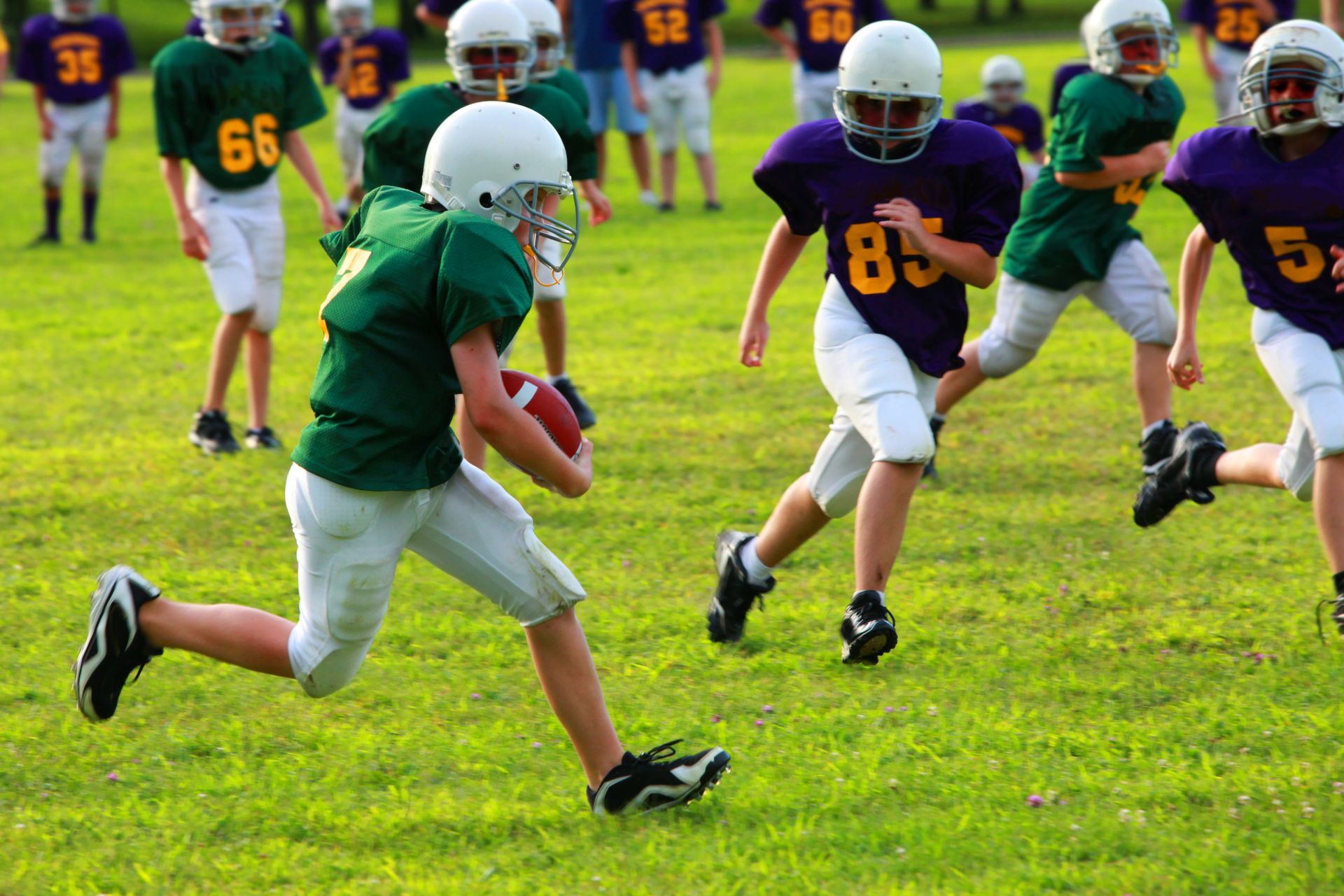A youth football player in a green jersey runs with the ball on a grass field as opponents in purple jerseys pursue.