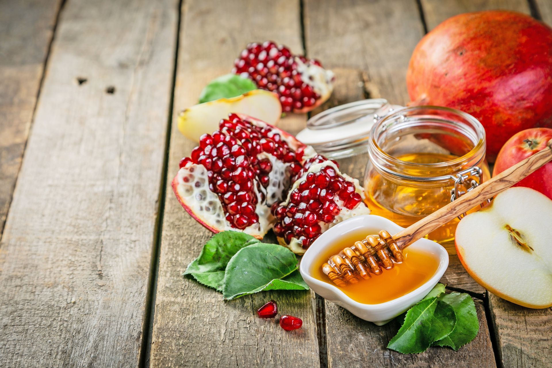 Pomegranate segments, apple slices, and honey in a jar and bowl with a honey dipper on a rustic wooden table.