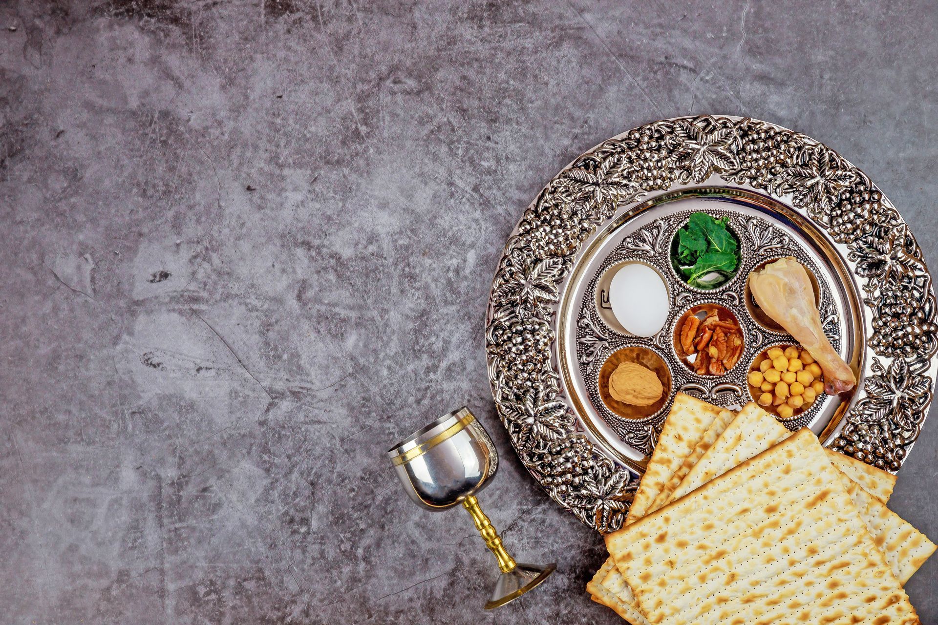 A Seder plate with six ceremonial foods, matzah, and a silver goblet on a gray, textured surface.