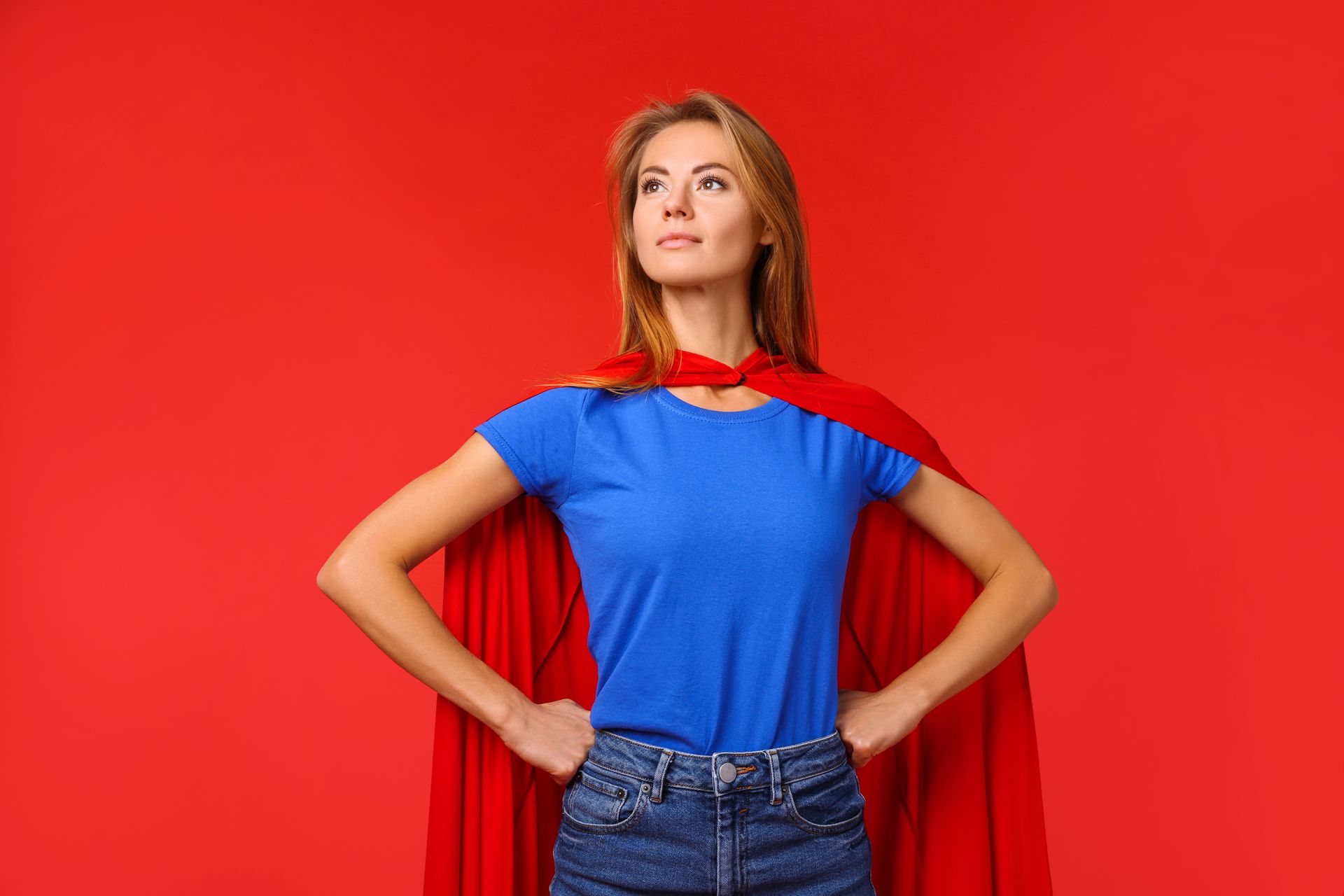 A person wearing a blue t-shirt and a red superhero cape stands with hands on hips against a vibrant red background.