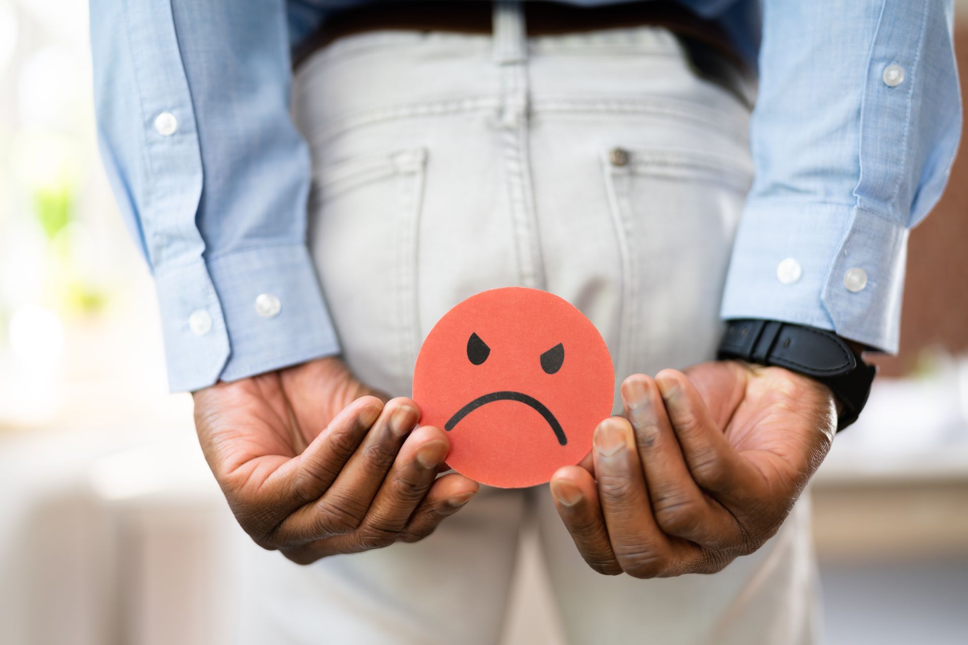 A person in light pants holds a red circle with an angry face expression over their backside.