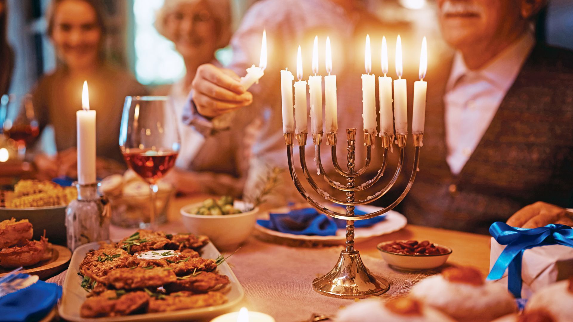 A hand lights a Hanukkah menorah on a table set with latkes and wine, surrounded by people at a festive celebration.