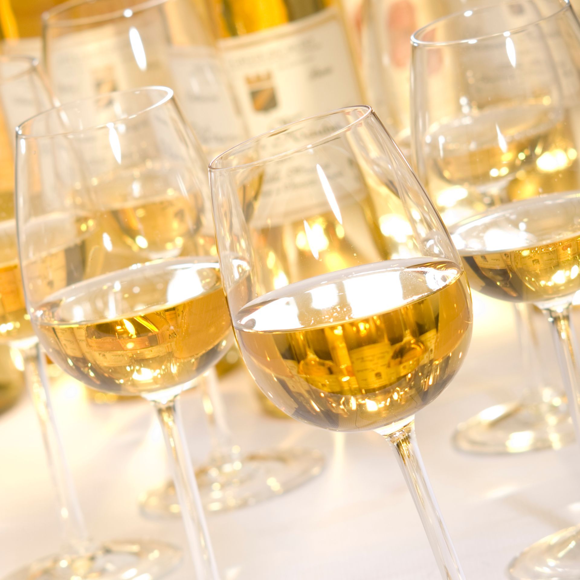 Several glasses filled with golden white wine are arranged on a white surface, with wine bottles blurred in the background.