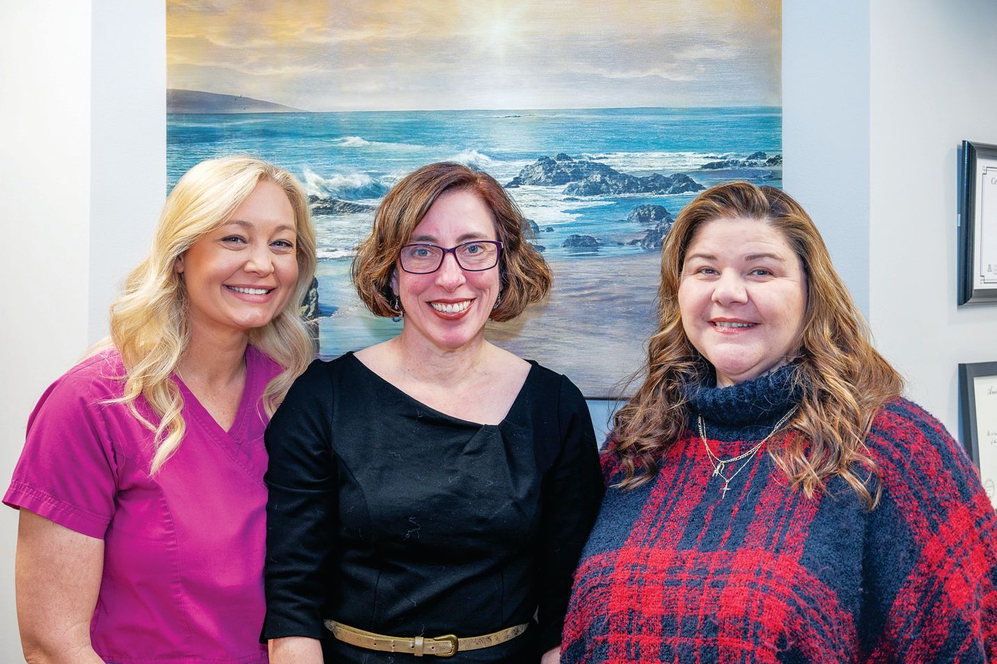 Three smiling individuals standing in front of a seascape painting inside an office.