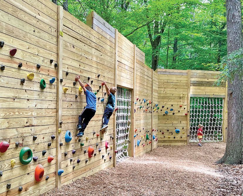 Two children climb a wooden wall with colorful rock grips at an outdoor park, while another child stands by a net barrier.