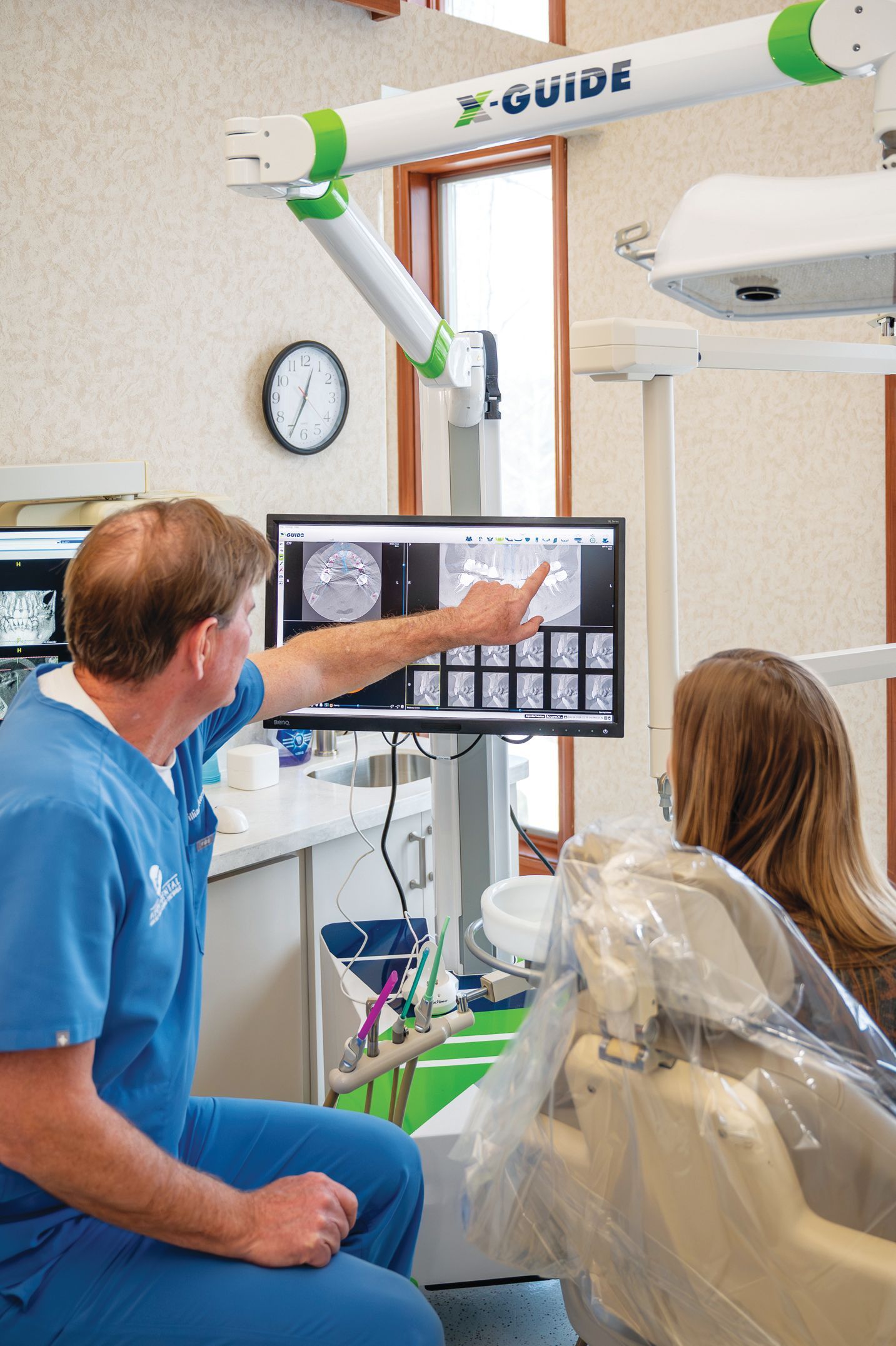 A dentist in blue scrubs points to dental imaging on a monitor while explaining a treatment plan to a seated patient.