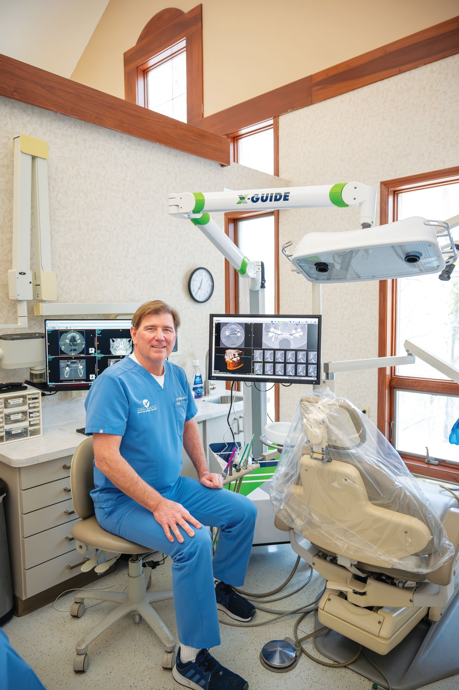 A smiling person in blue scrubs sits in a dental office next to a patient chair and medical computer monitors.
