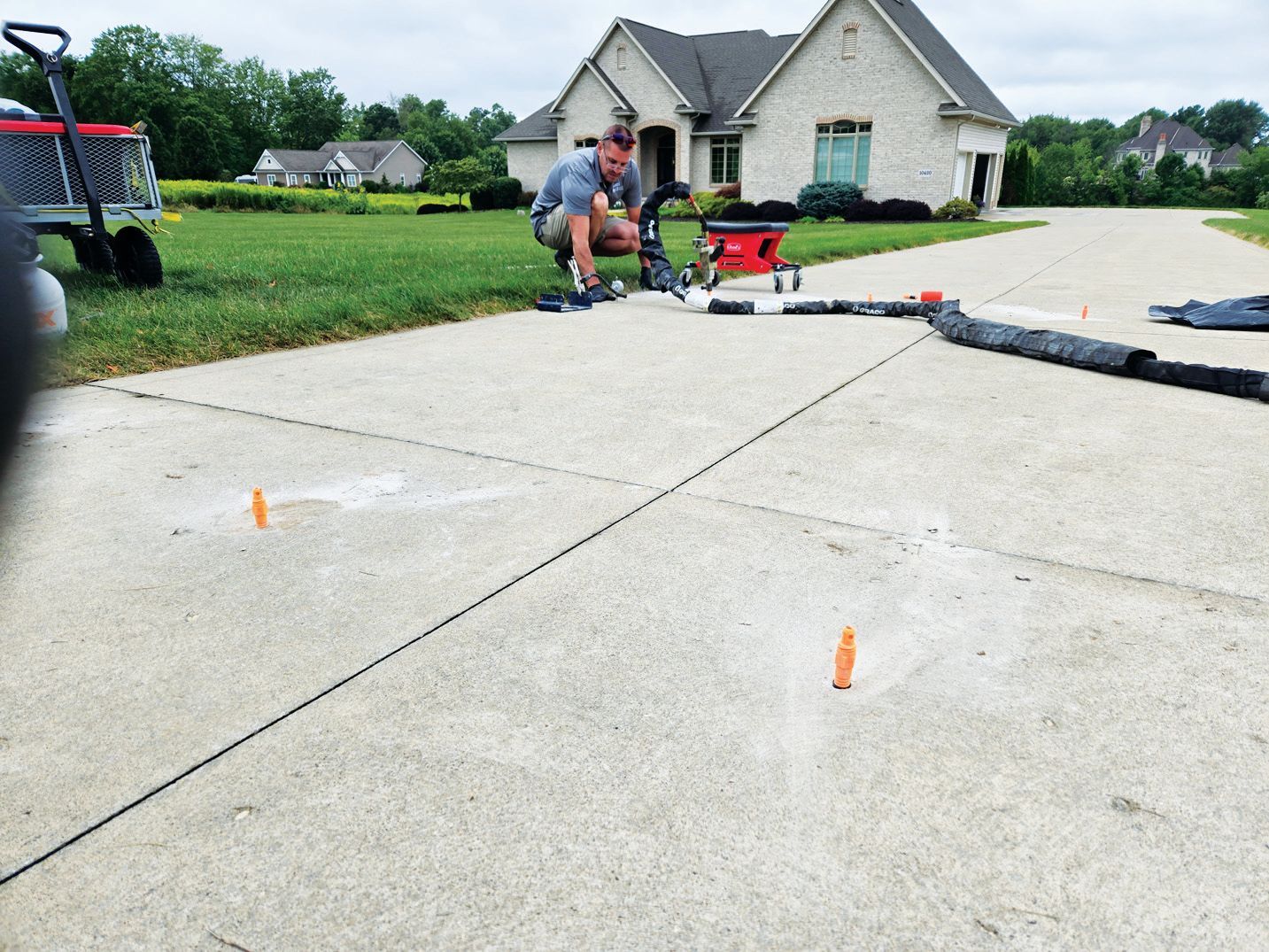 A worker kneels on a concrete driveway, preparing equipment for repair, marked by orange cones near a suburban house.