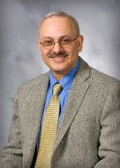 Professional headshot of a person wearing glasses, a blue striped dress shirt, a patterned tie, and a gray textured blazer.