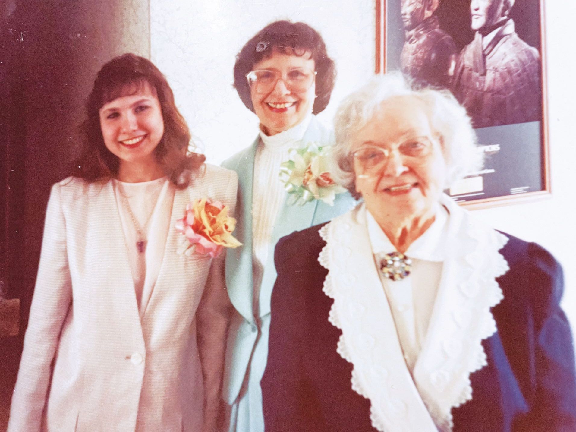 Three people in formal attire pose together indoors, with a framed portrait hanging on the wall in the background.
