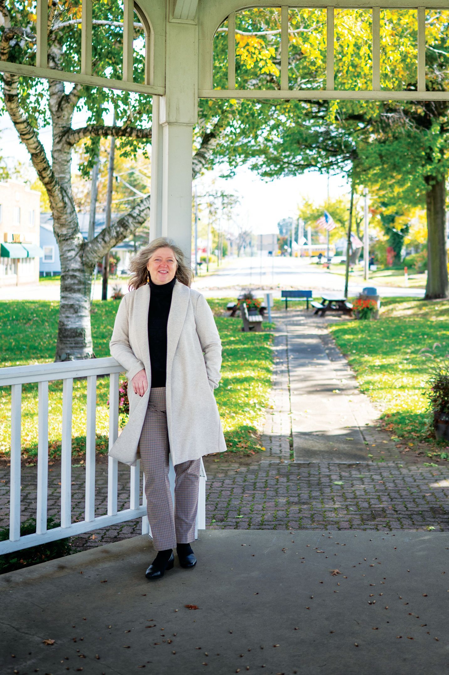 A person in a cream coat and patterned pants stands on a gazebo porch overlooking a sunny park pathway.