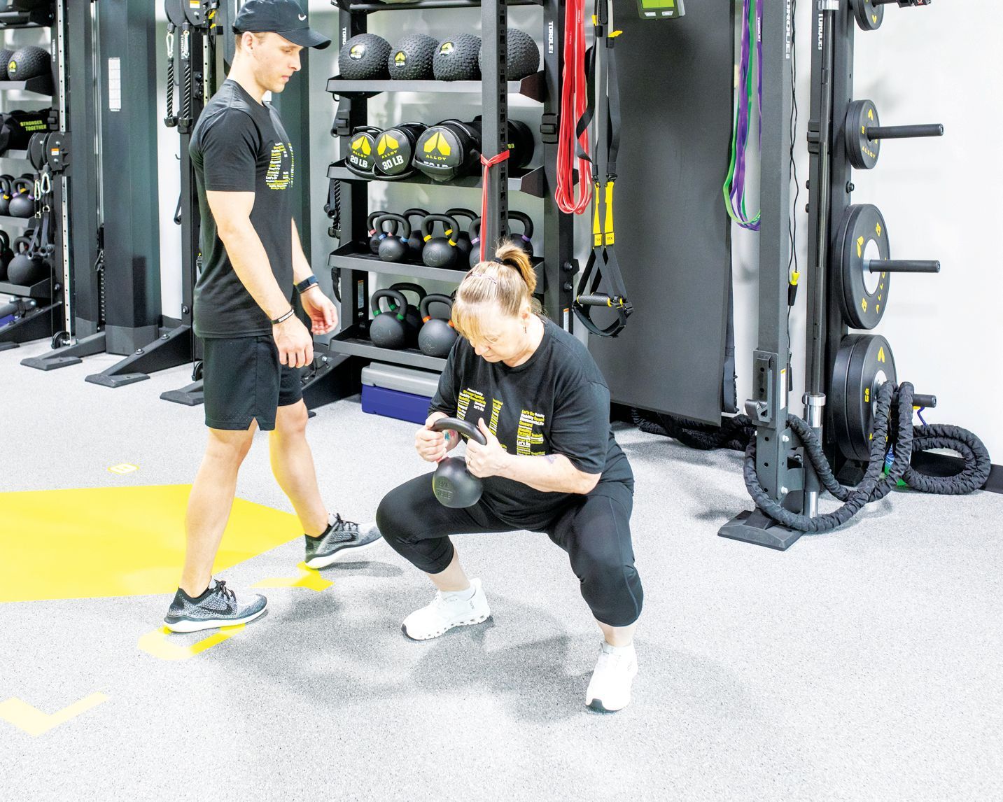 A personal trainer watches as a person performs a kettlebell goblet squat in a gym with exercise equipment in the background.