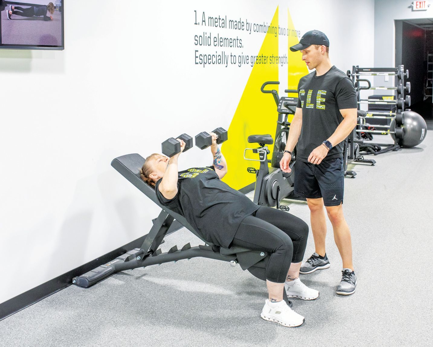 A person performs an incline dumbbell chest press on a bench in a gym as a trainer stands nearby supervising.