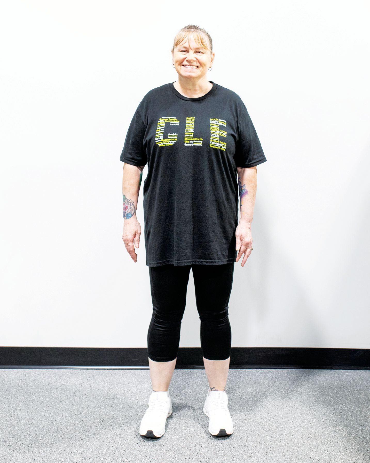 A person stands smiling in front of a white wall, wearing a black graphic t-shirt, black leggings, and white sneakers.