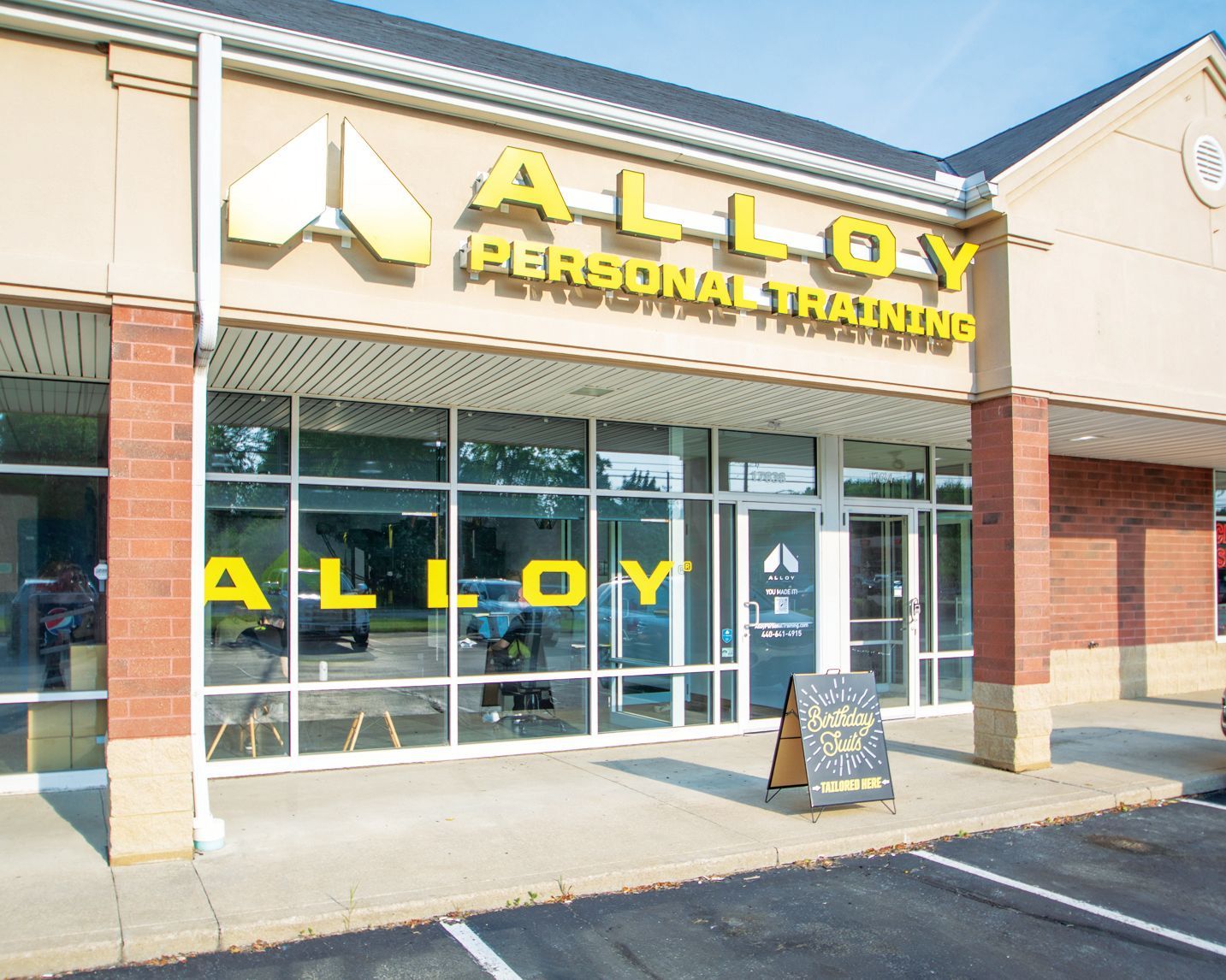 The storefront for Alloy Personal Training, featuring yellow signage on a brick and beige building with large windows.
