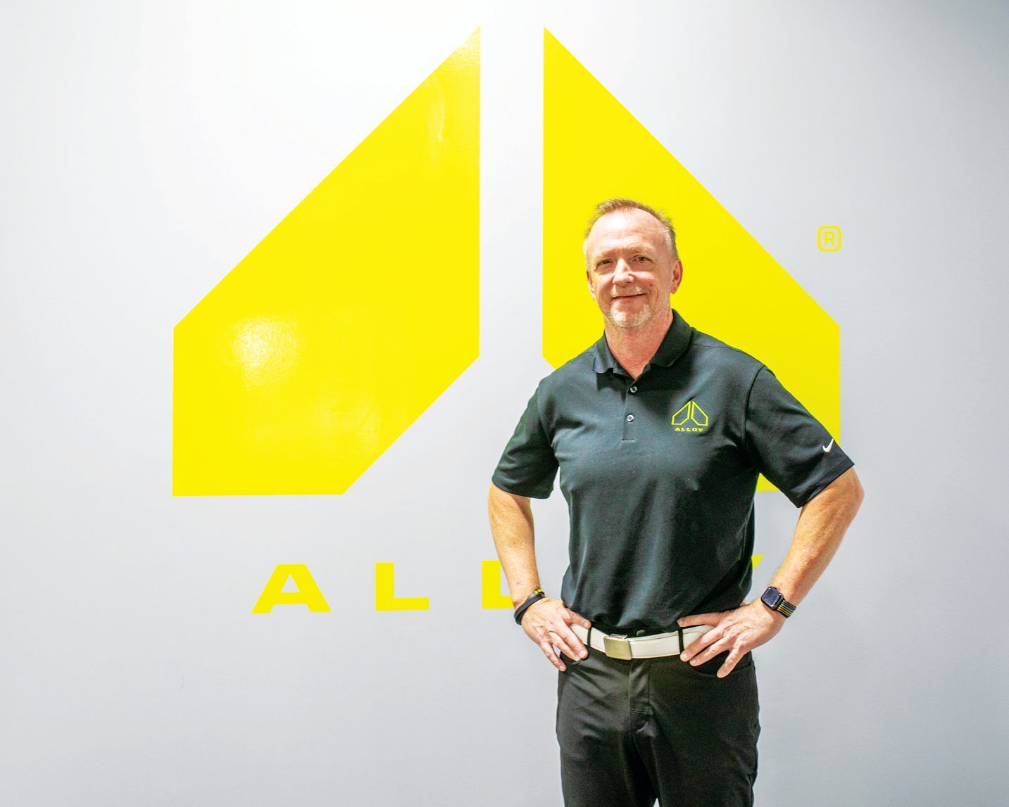A person stands with hands on hips against a light grey wall featuring a large, yellow geometric logo.