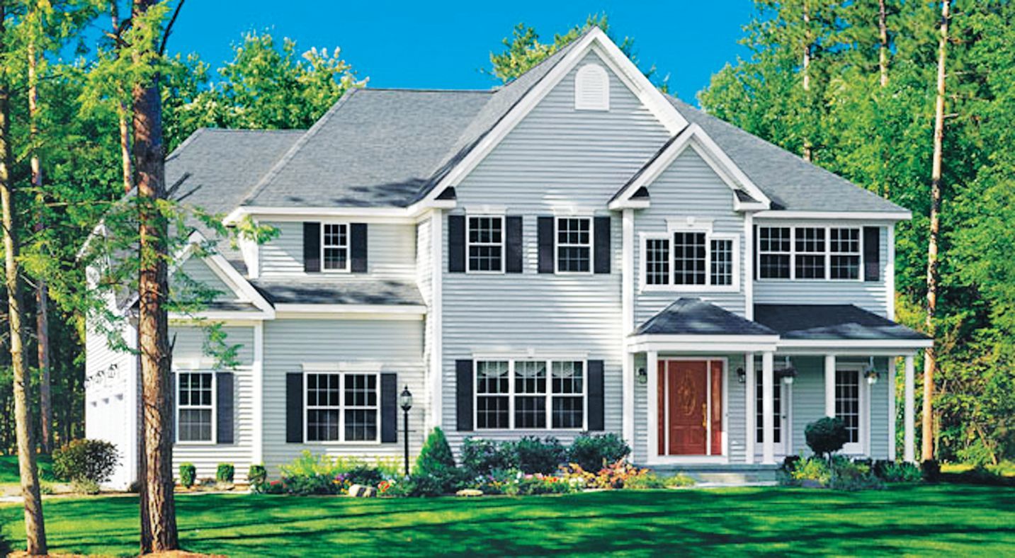 A two-story light gray house with black shutters and a red front door, surrounded by green trees and a grassy lawn.