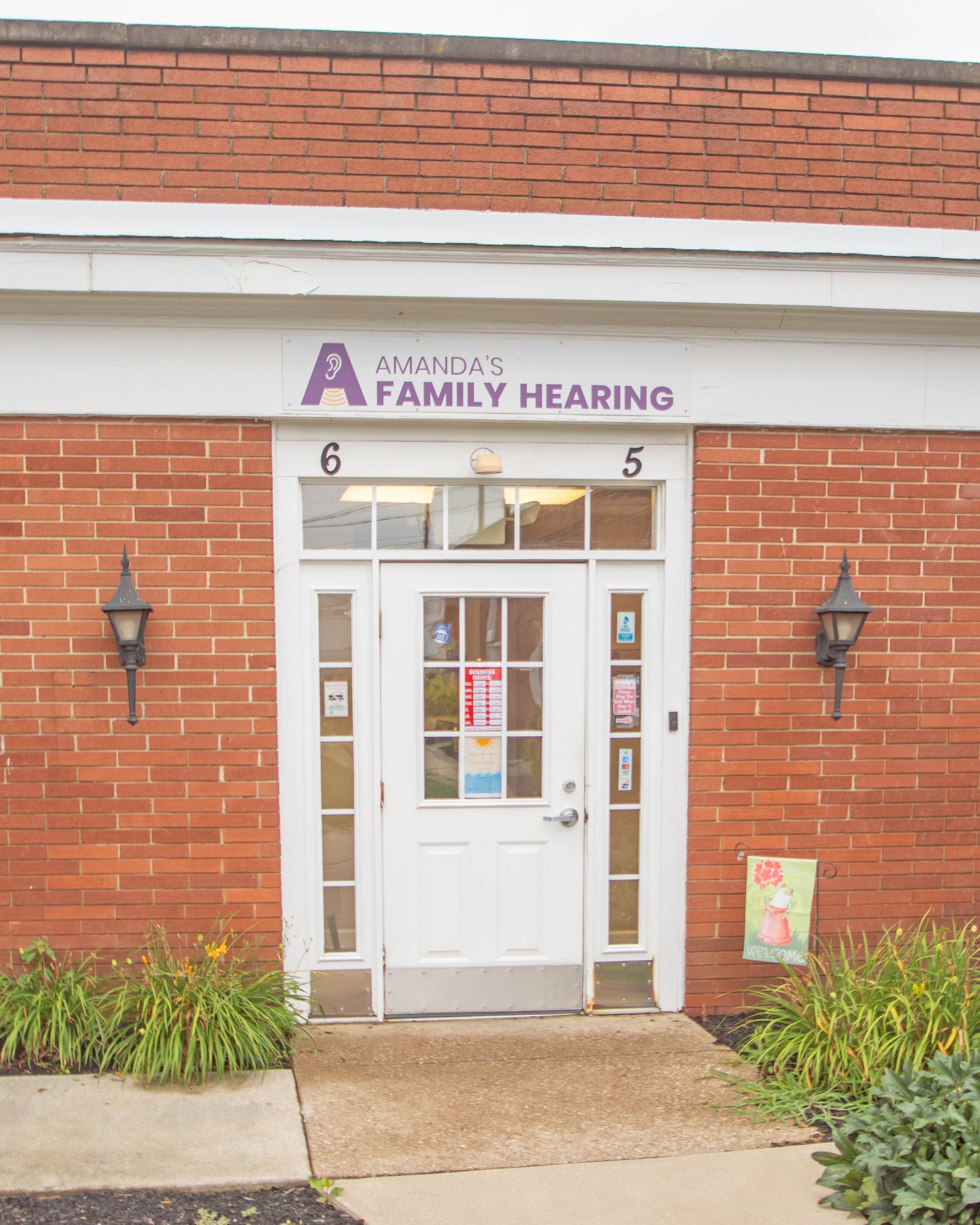 The red brick storefront of American Family Hearing with a white front door, hanging lanterns, and green shrubs.